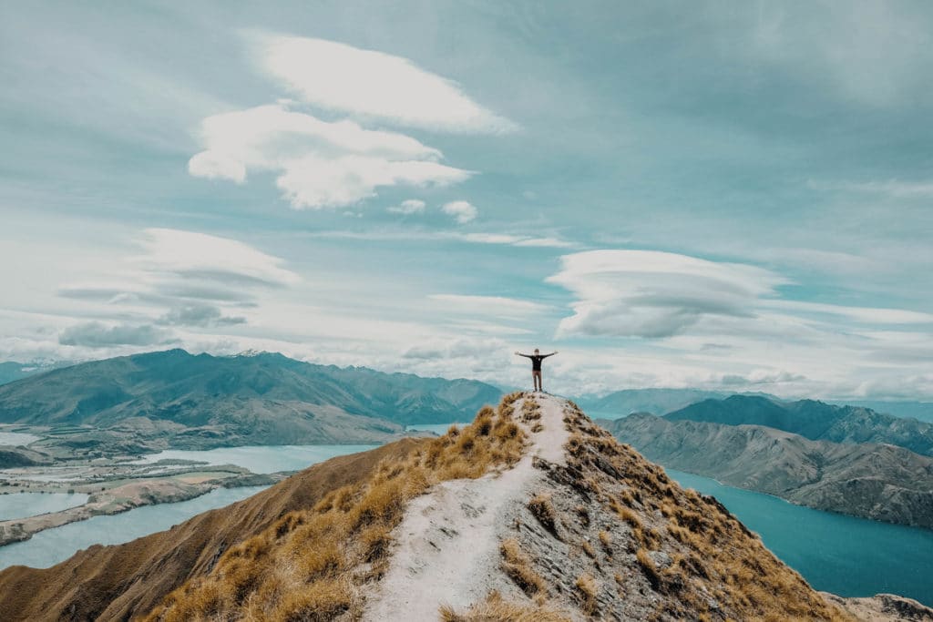 Backpacker en haut d'une montagne en Nouvelle Zélande qui écarte les bras devant le paysage de carte postale